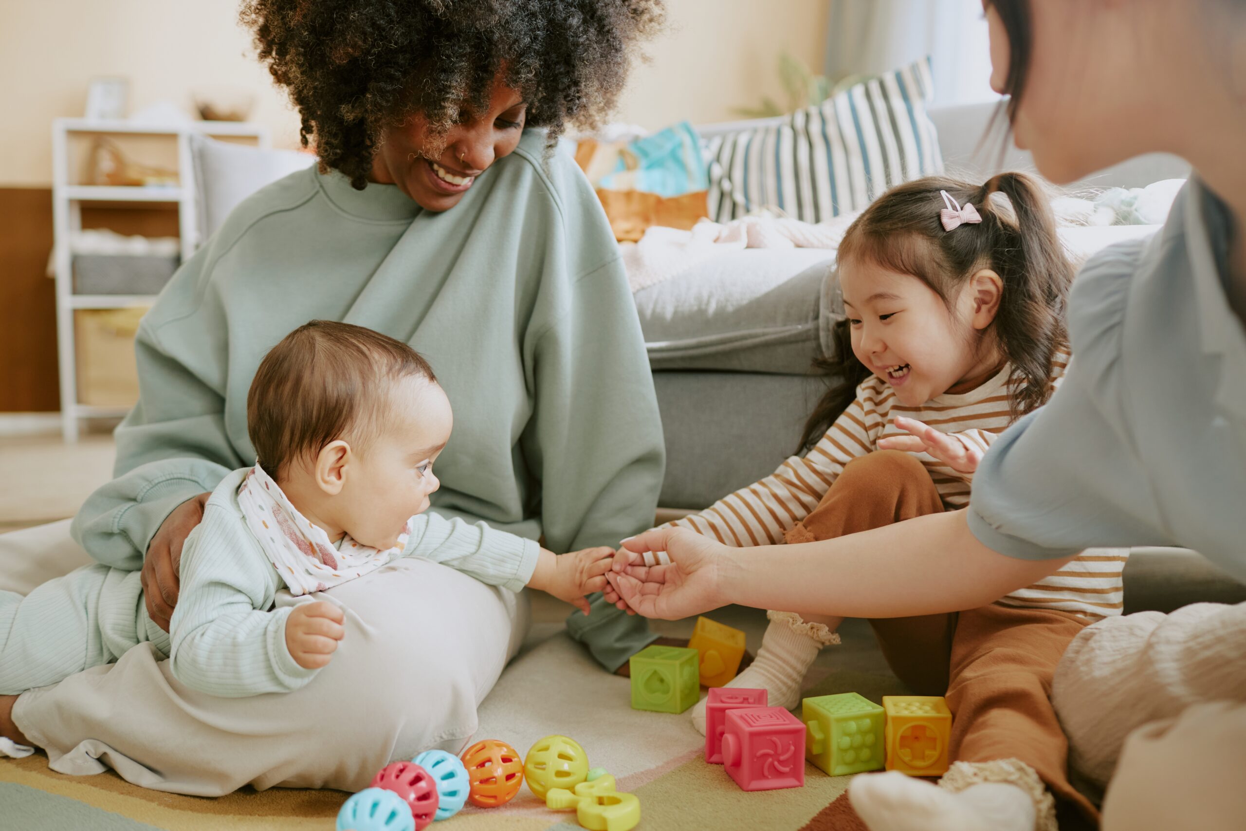 Baby boy touching hands of Asian girl and her mother while his African American mom watching them and smiling